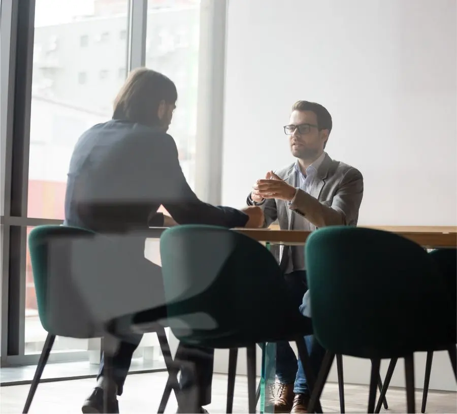 Two men are sitting at a table in an office having a conversation.