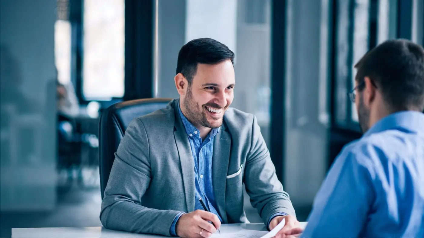 Two men are sitting at a table in an office having a conversation.