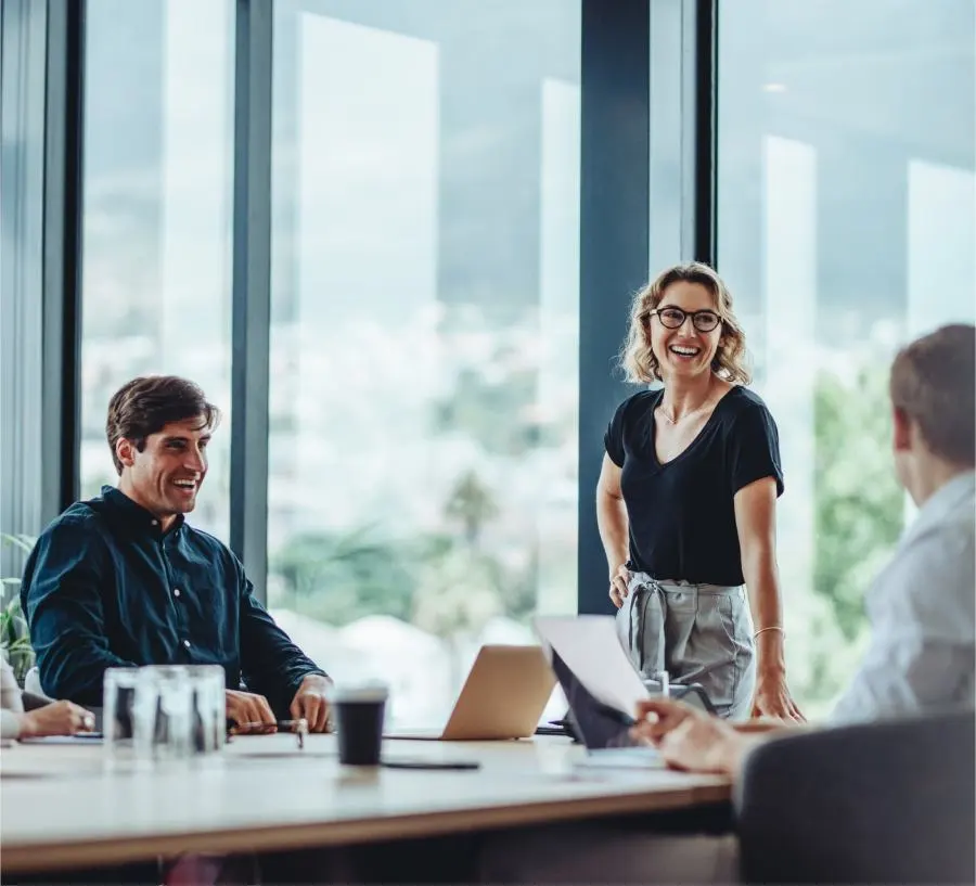 Picture of a standing laughing woman and a seated laughing man during a meeting in the office with a view of the greenery.