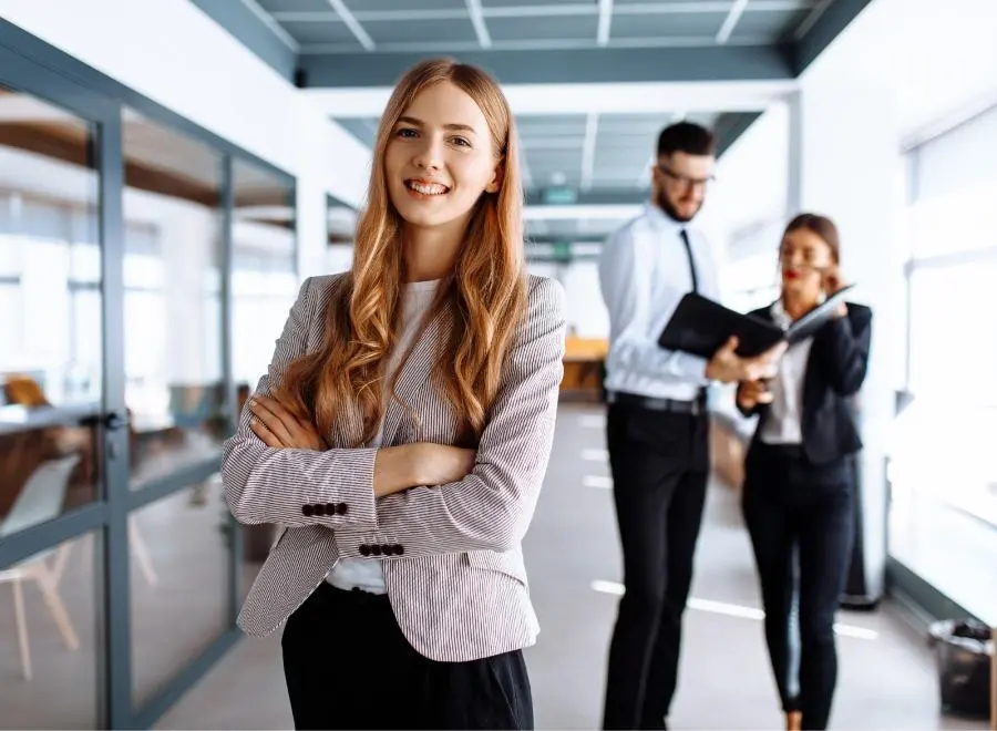 A young woman in the office laughs confidently into the camera with her arms crossed.