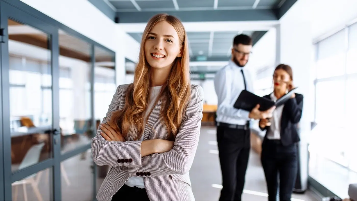 A young woman in the office laughs confidently into the camera with her arms crossed.