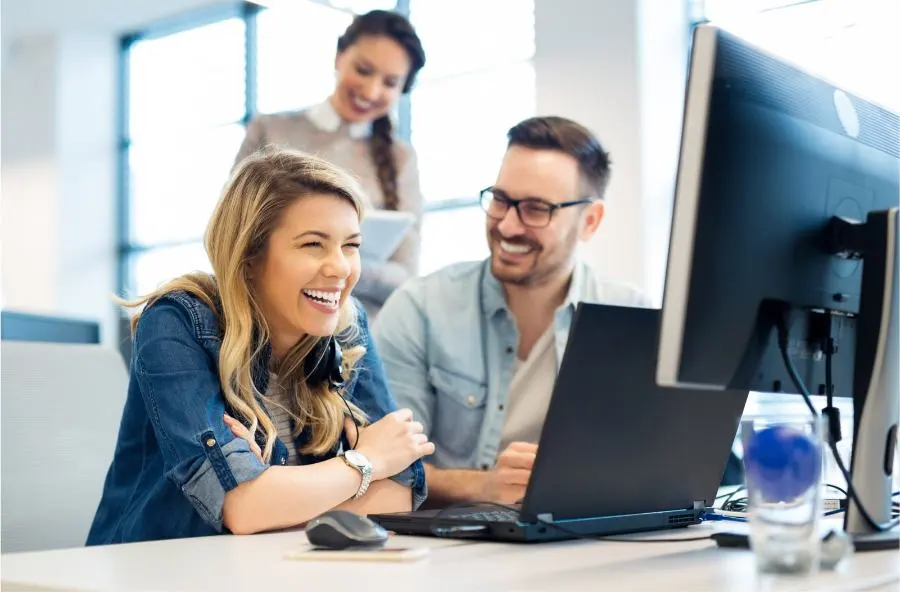 A woman and a man sit laughing in front of a computer.