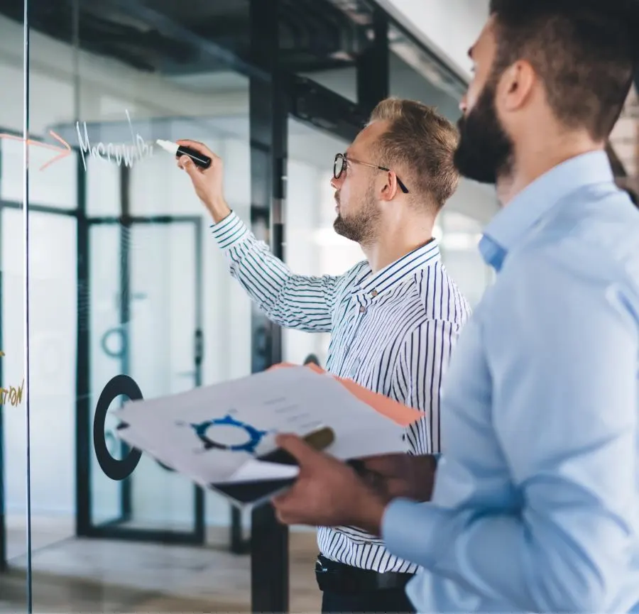 Two men are taking meeting notes on a glass wall looking concentrated.